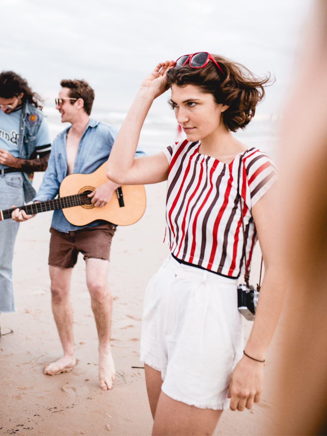 Woman in striped shirt and white shorts standing on a beach with a man playing guitar in the background.