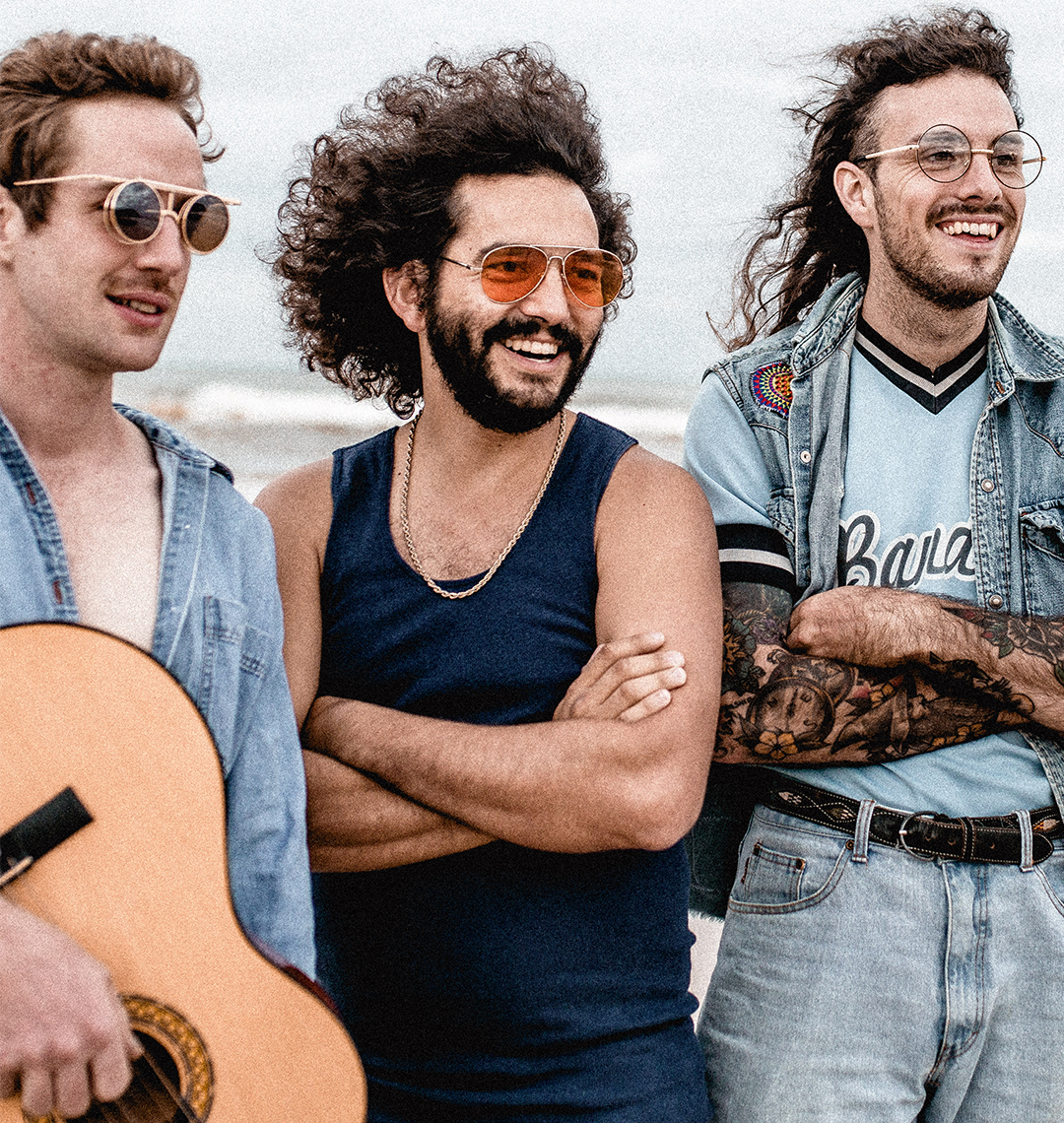 Three men standing together outdoors, two of whom are holding guitars.