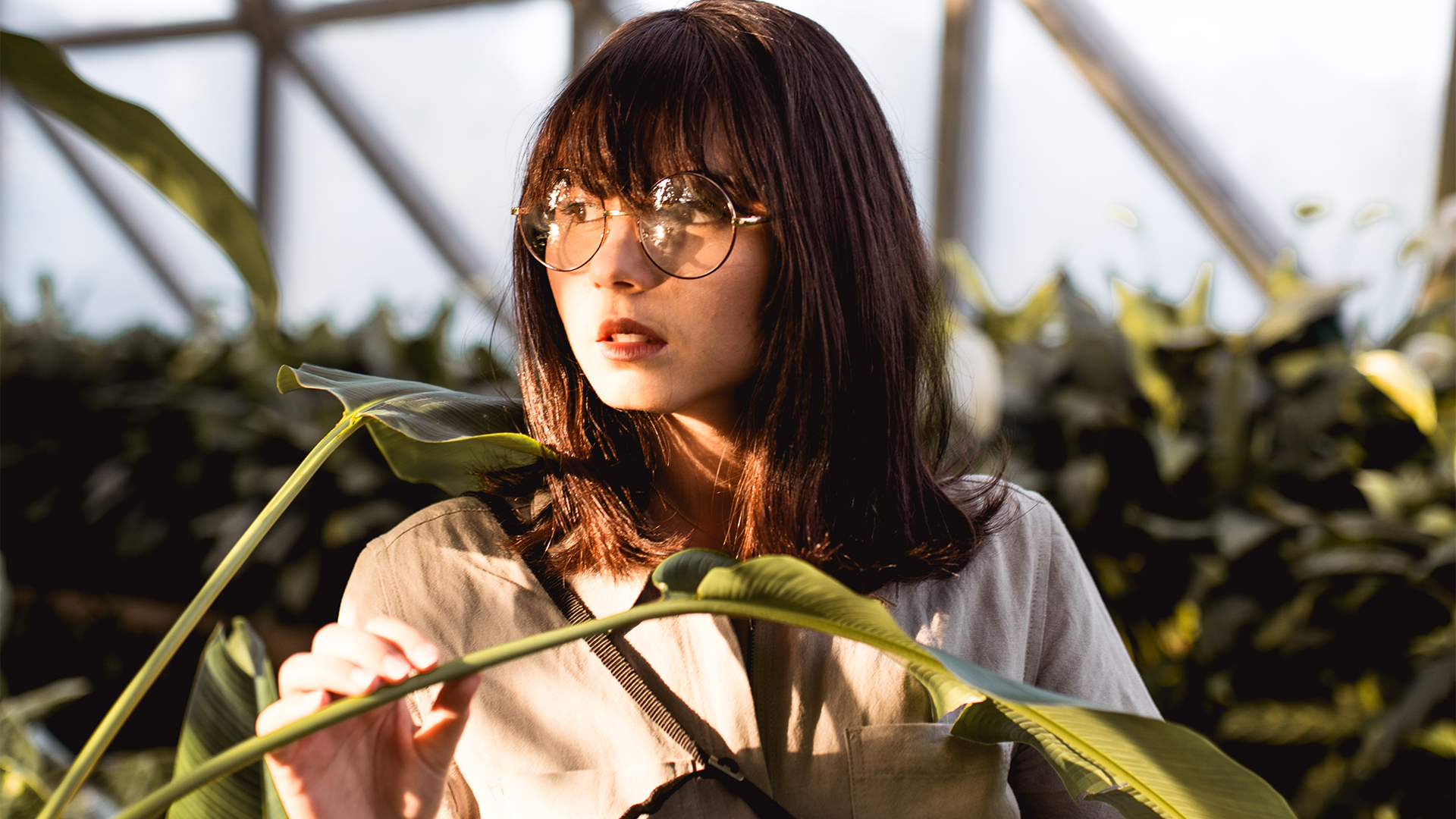 Woman wearing glasses and a light-colored shirt, standing among green plants.