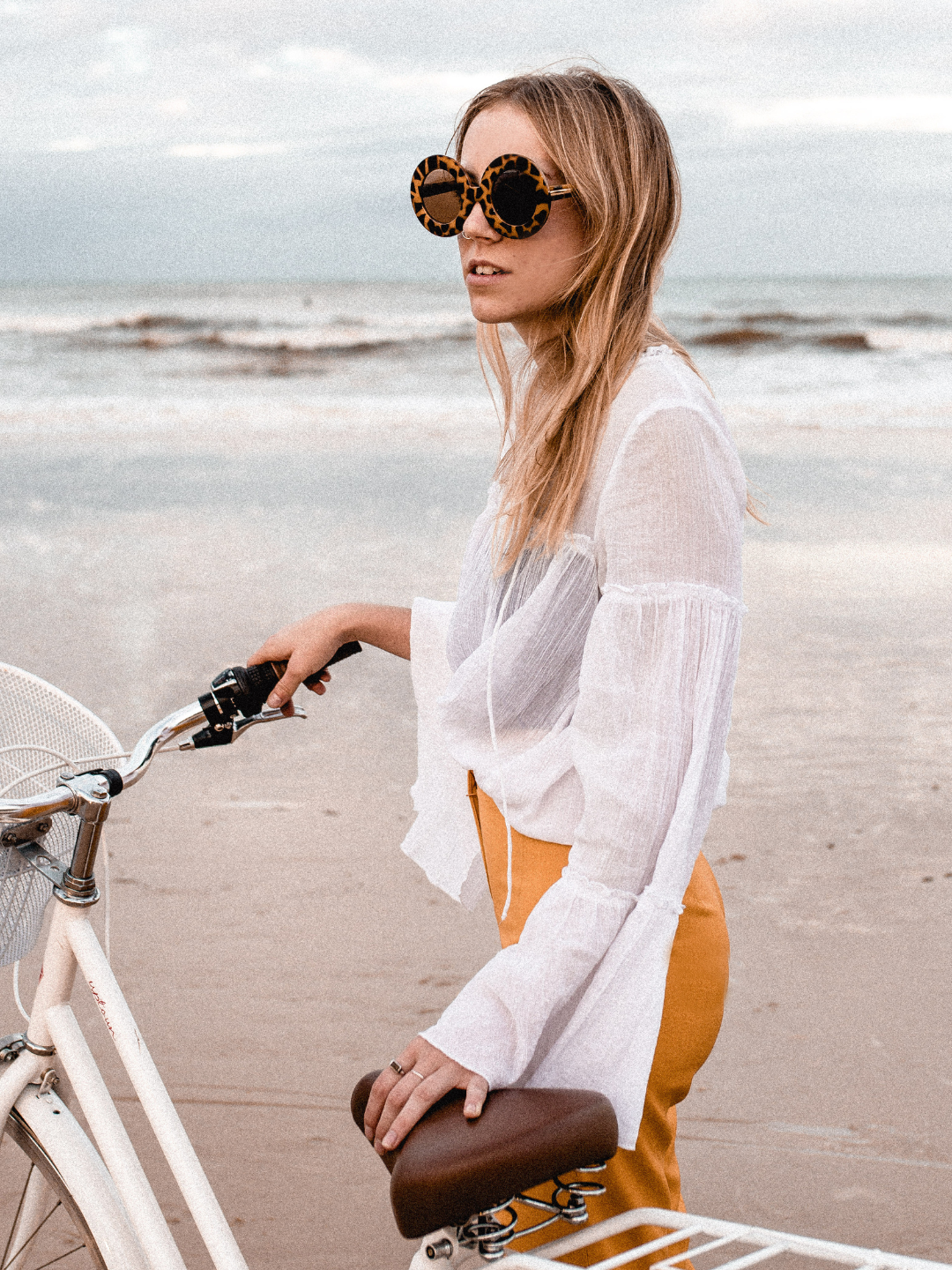 Woman with sunglasses and a white shirt standing next to a bicycle on a beach.