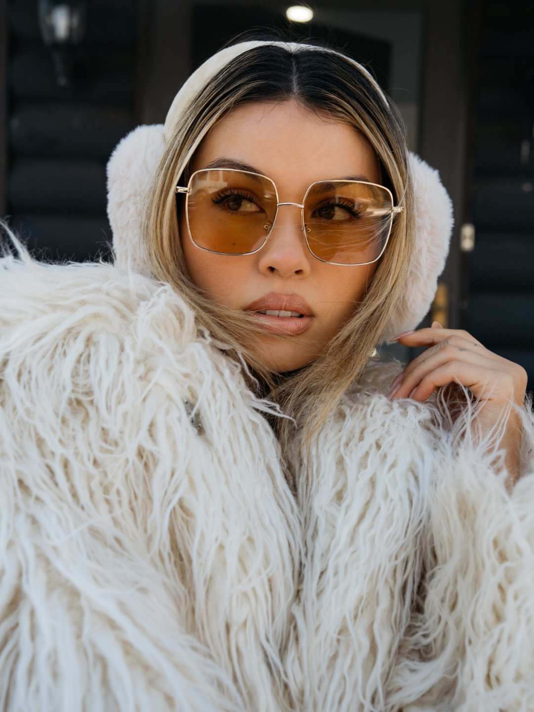 Woman wearing a white fur coat and sunglasses indoors.