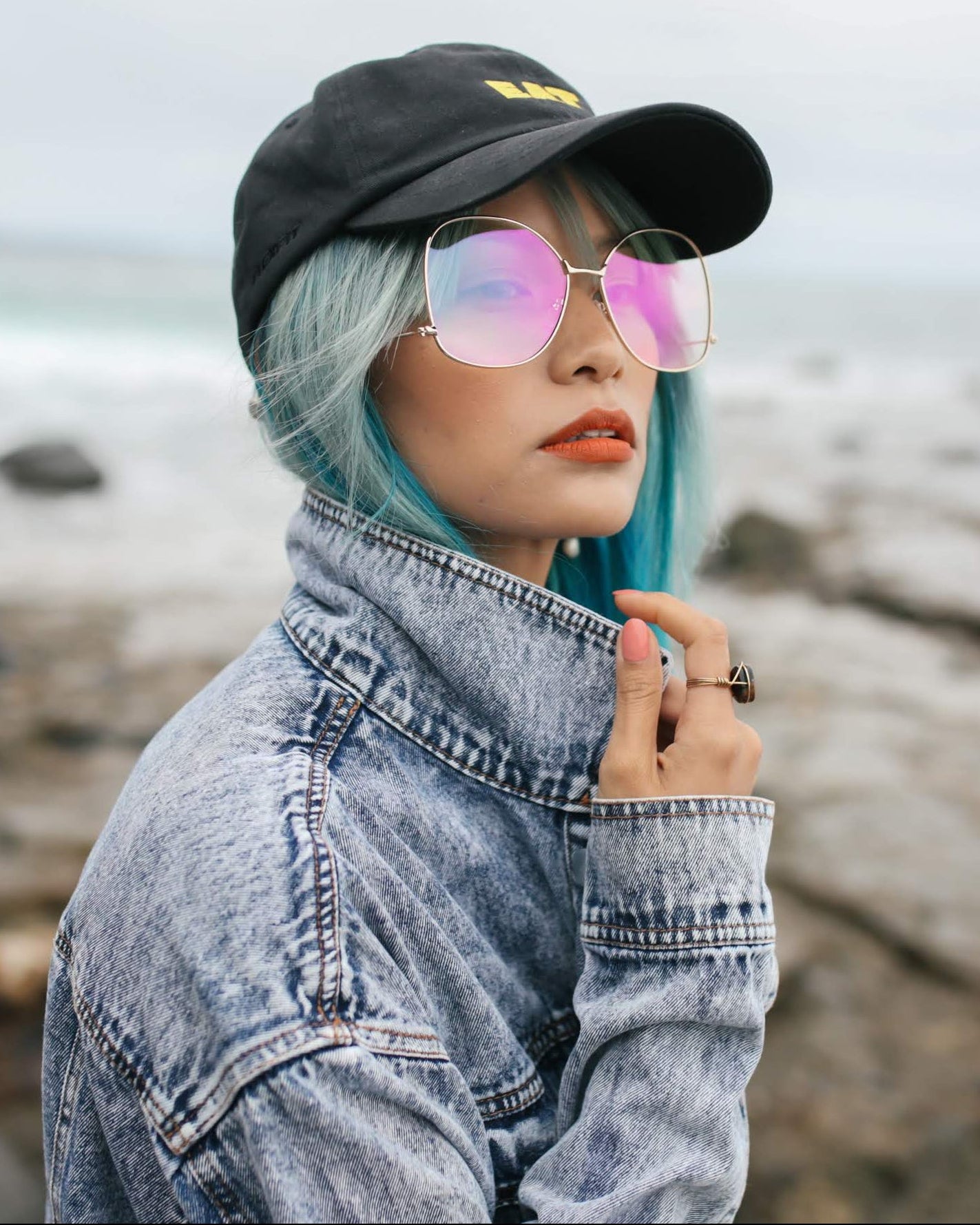 Person with teal hair and pink sunglasses standing on a beach.