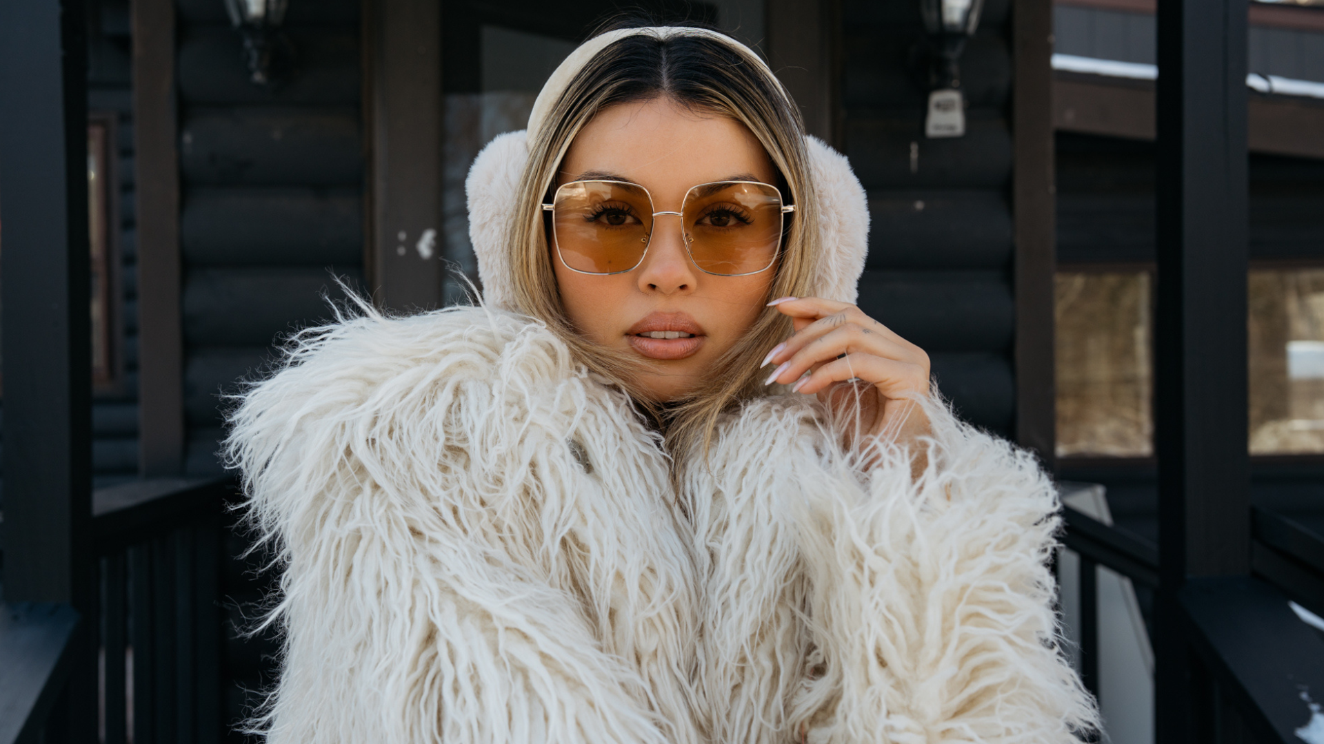 Woman wearing a white fur coat and sunglasses in front of a wooden wall.