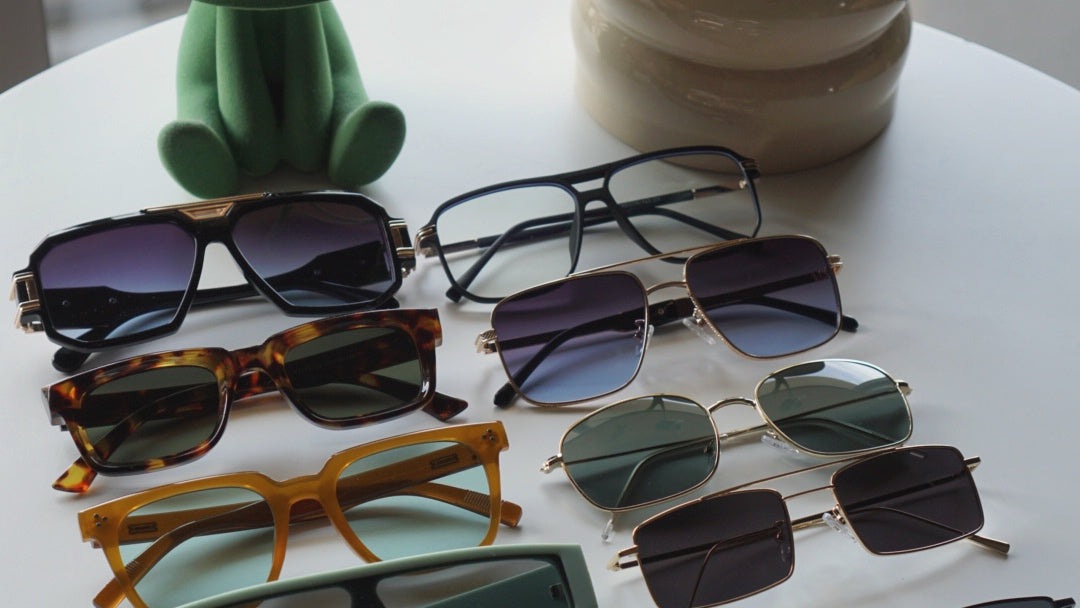 Collection of sunglasses on a white surface with a green plush toy and beige container in the background.