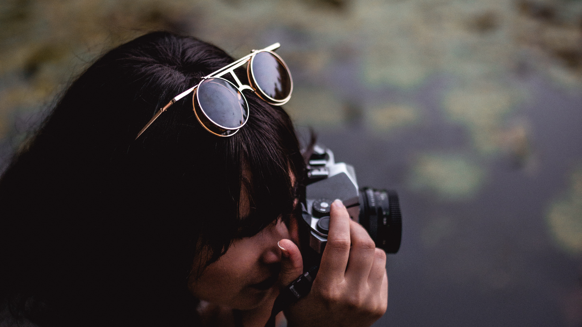 Person holding a camera with sunglasses on, blurred natural background