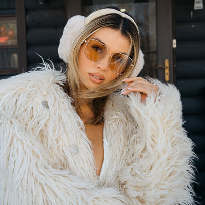 Woman wearing a white fur coat and sunglasses in front of a log cabin.