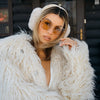 Woman wearing a white fur coat and sunglasses in front of a log cabin.